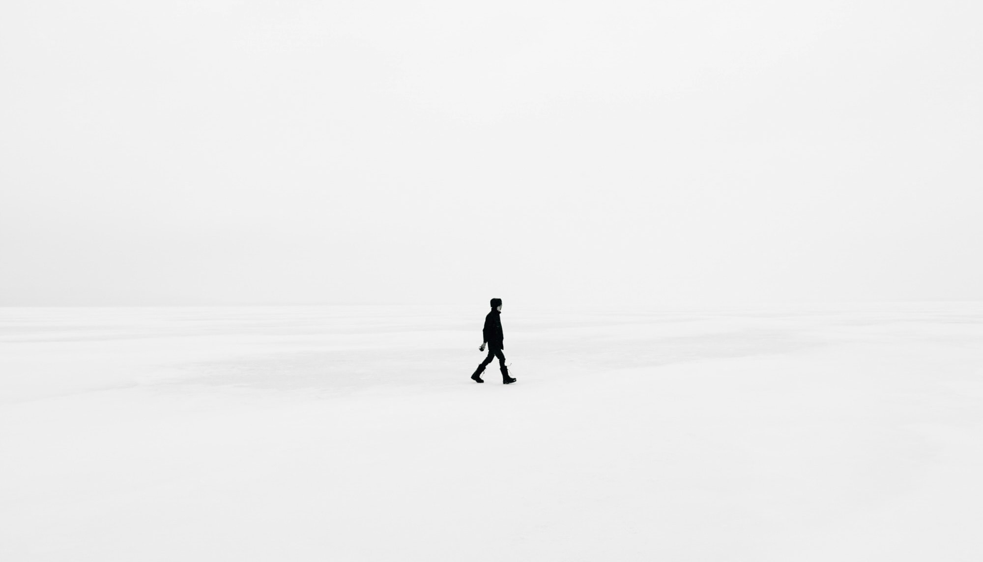A black and white photograph of a lone figure walking across a stark white landscape